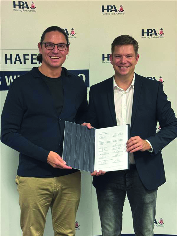 Zwei Männer stehen vor einer Fotowand der Hamburg Port Authority (HPA) und halten einen unterzeichneten Vertrag in den Händen.
