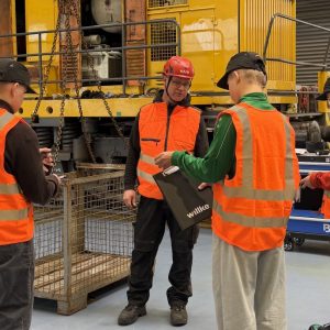 Mehrere Personen in orangefarbenen Warnwesten stehen in einer großen Industriehalle vor einem gelben Schienenfahrzeug und tauschen sich aus.