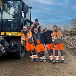 Fünf Gleisbauarbeiter in orangefarbener Schutzkleidung stehen neben einem Schienenfahrzeug auf einer Schienenbaustelle.