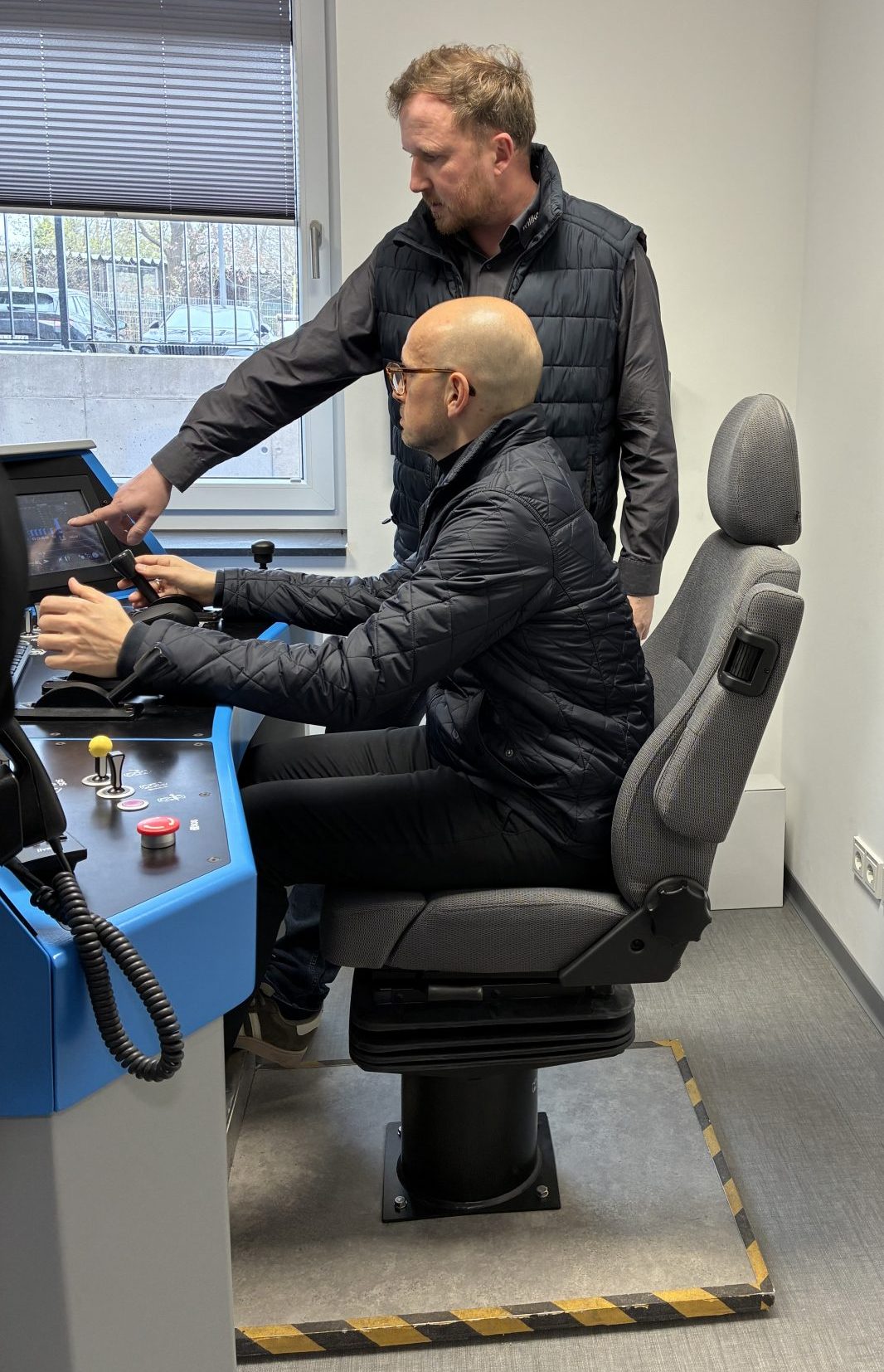Zwei Männer bedienen zusammen einen Eisenbahnsimulator, wobei einer sitzt und der andere ihm etwas am Bildschirm erklärt.